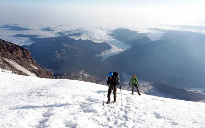 two people wearing mountaineering gear stand on a very high snowy field overlooking a vast mountainous landscape